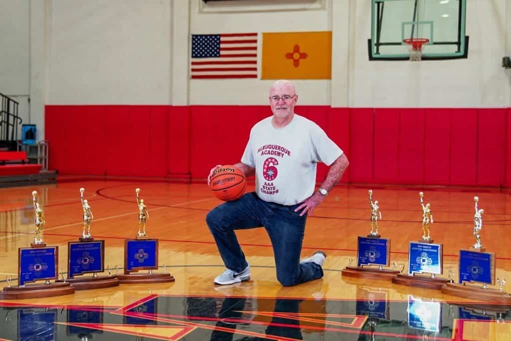 Albuquerque Academy coach kneeling with basketball trophies on gym floor.