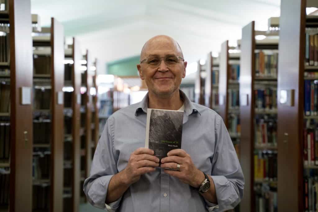 Adult male smiling in library holding a book about nature and survival.