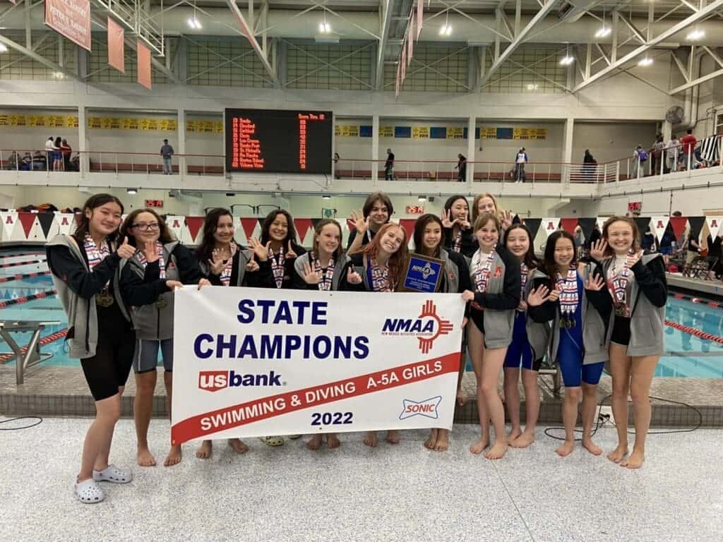 Girls swimming team celebrating state championship victory at Albuquerque Academy indoor pool.