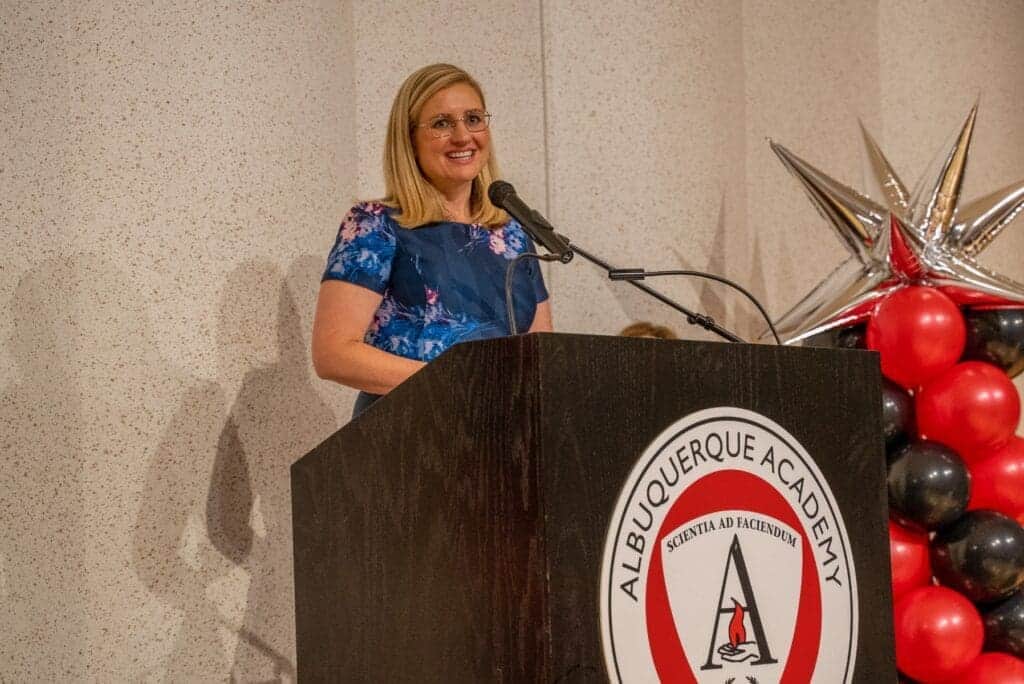 Elegant woman speaking at Albuquerque Academy graduation ceremony with school logo and decorations.