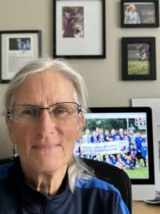 A woman wearing glasses in an office with framed sports and school photos and a computer screen displaying a group photo.