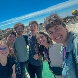 A diverse group of students smiling together outdoors with Albuquerque city landmarks in the background.