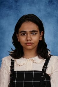 Young girl with dark hair and makeup, wearing a white blouse and black checkered dress against a blue background.