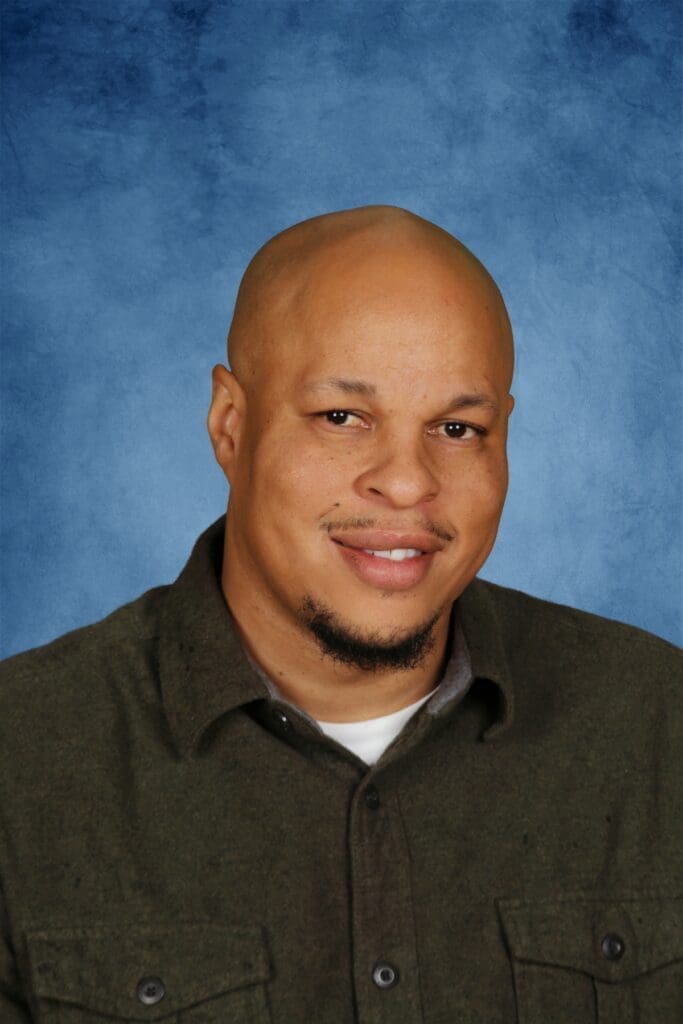 Bald man with goatee smiling against blue background at Albuquerque Academy.
