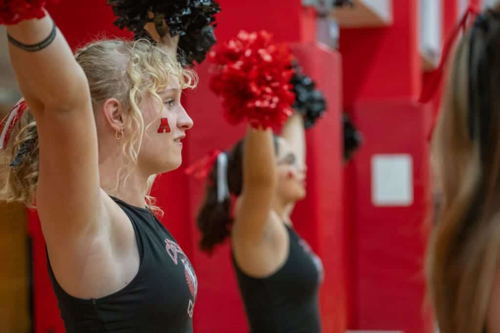 Cheerleaders practicing routine at Albuquerque Academy in a gym with red walls and pom-poms.