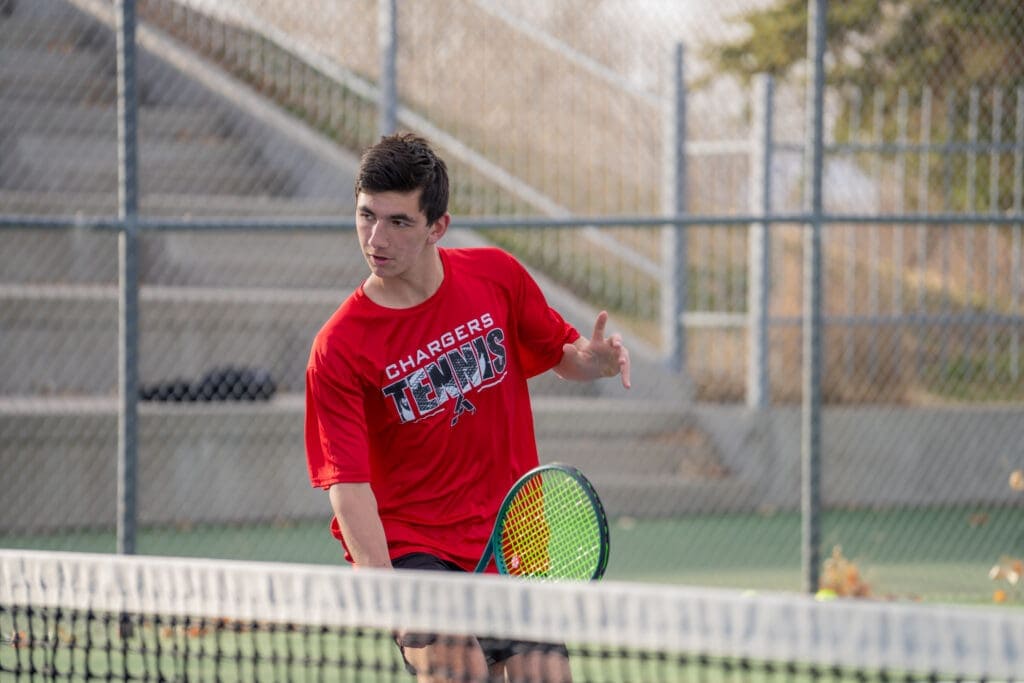 Young male tennis player practicing on an outdoor court, wearing a red Albuquerque Academy Chargers shirt.