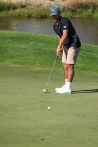 A young man practicing golf on a lush green course, wearing casual attire and a cap, with a pond in the background.