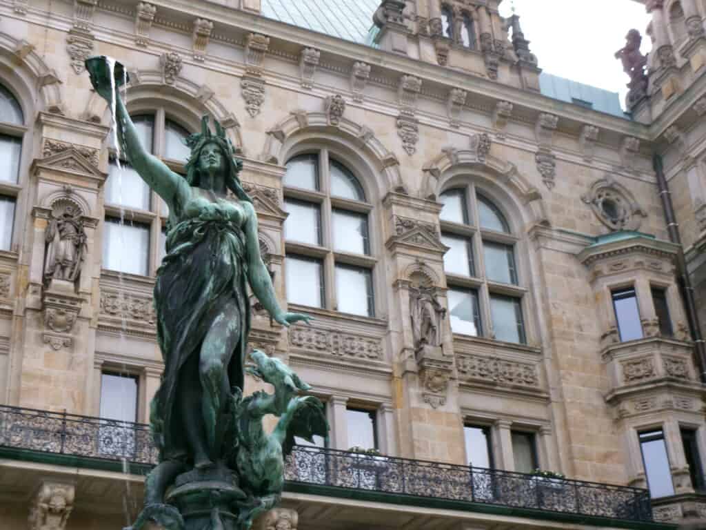 Bronze statue of a woman with flowing hair and draped clothing on a fountain, historic building in background.