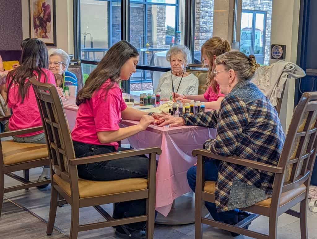 A young girl and senior women painting nails at a community center table with elderly women in the b.