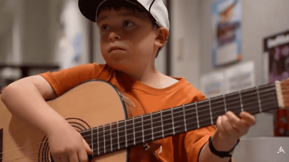 A young boy playing an acoustic guitar in a classroom setting, promoting summer learning activities.