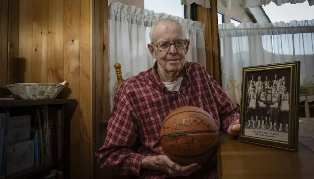 A senior man holding a basketball with a framed team photo on a wooden table at Albuquerque Academy.