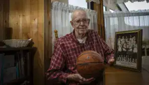 A senior man holding a basketball with a framed team photo on a wooden table at Albuquerque Academy.