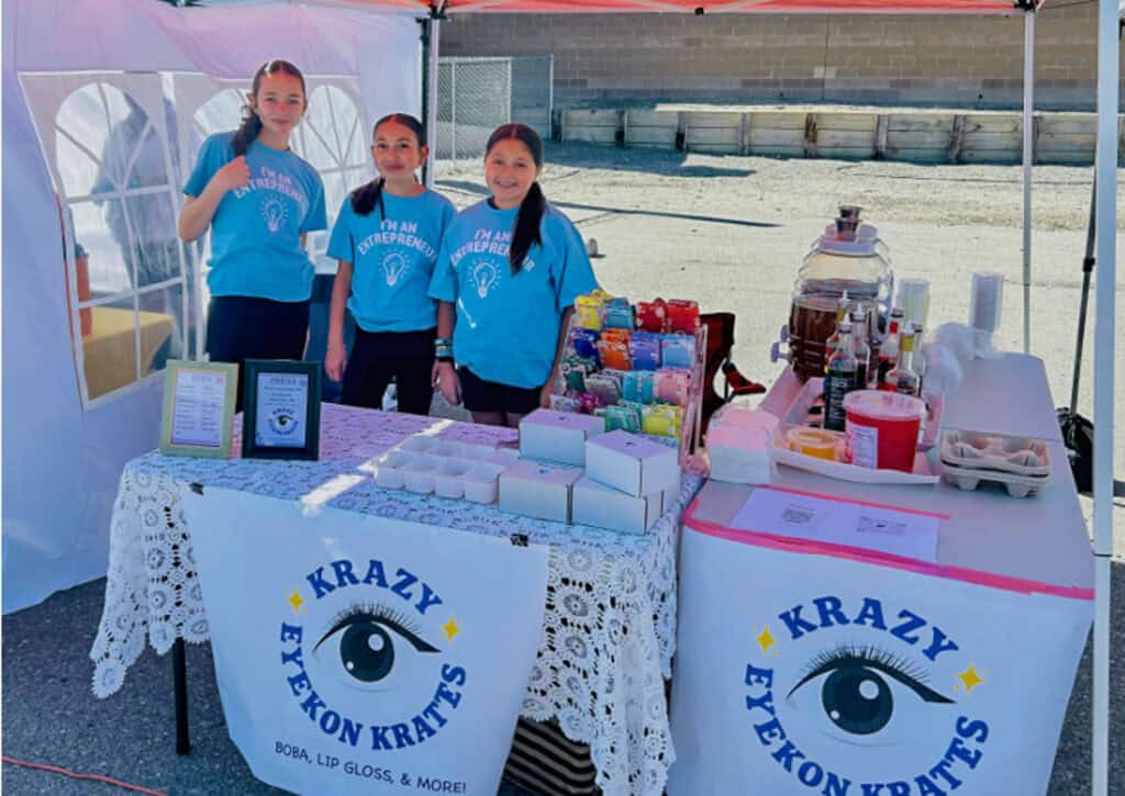 Three girls at Krazy Eyekon Krates booth selling crafts and baked goods.