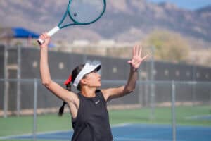 Albuquerque Academy girl tennis player serving on outdoor court during match.