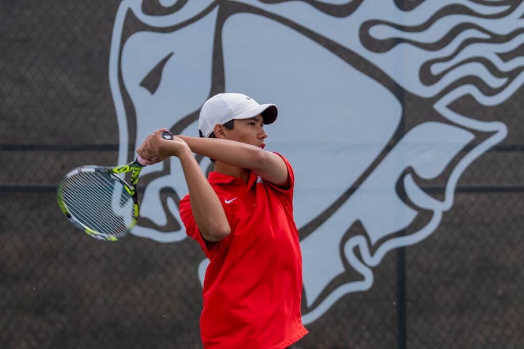 Albuquerque Academy student playing tennis on outdoor court with school logo background.