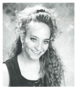 A woman with curly hair smiling, wearing a black top, in black and white photo.