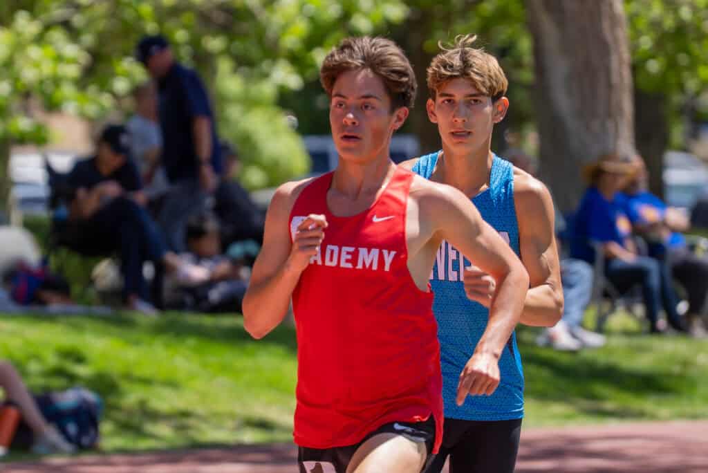 A young male runner in a red Albuquerque Academy track uniform competing in a race outdoors.