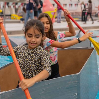 Two girls paddling a cardboard boat in a swimming pool during a school event at Albuquerque Academy.