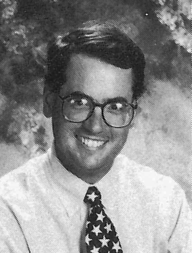 Black and white portrait of a young man with glasses, smiling, wearing a star-patterned tie.