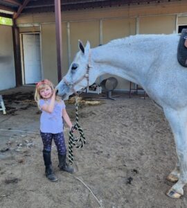 Young girl with blonde hair petting a white horse in a stable yard.