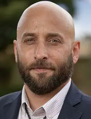 Close-up of a bald man with a beard in a suit outdoors.
