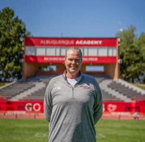 Albuquerque Academy coach standing on athletic field with stadium in background.