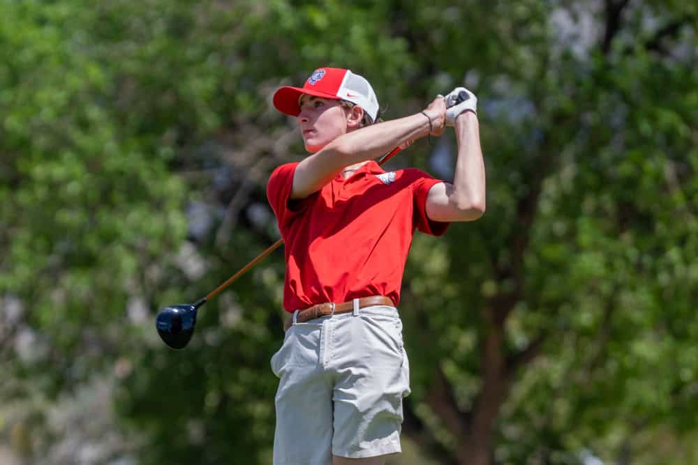 Young golfer in red shirt and cap swings club on golf course.