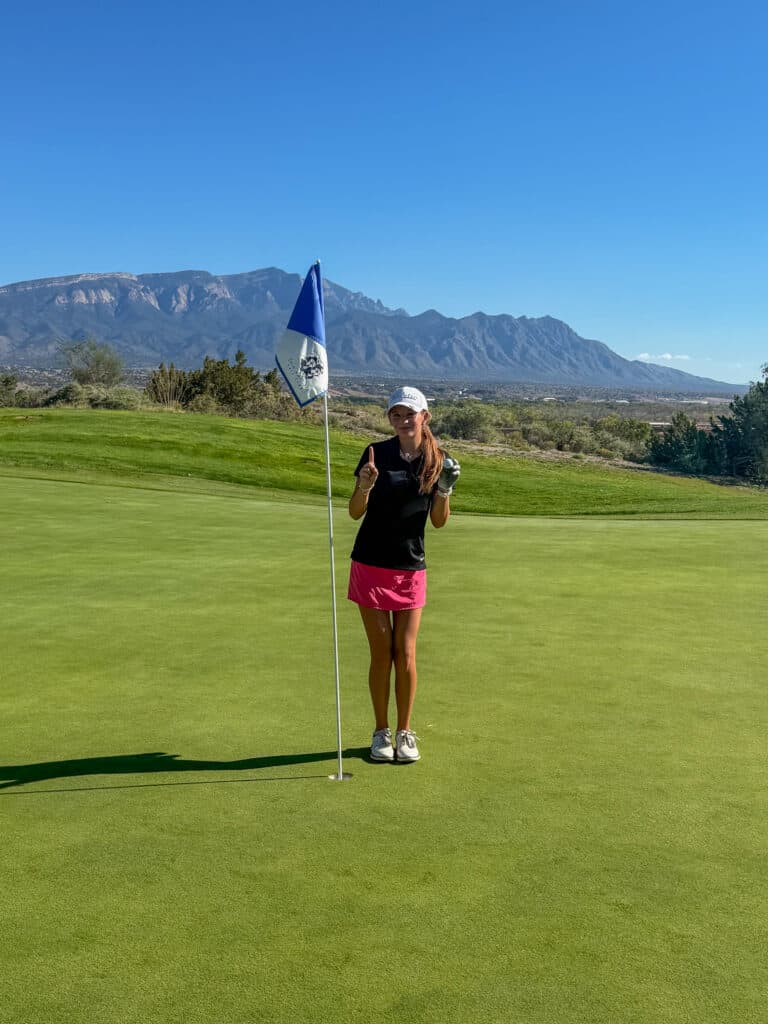Young woman celebrating a hole-in-one on a golf course with mountains in the background.